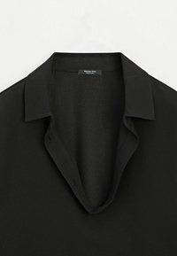 Chemise noire à col avec encolure ouverte montrant la partie haute de l’avant et une petite étiquette de marque à l’intérieur du col.