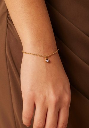 Un bracelet en chaîne dorée présente un petit pendentif en pierre précieuse violette, conçu avec un motif de mailles délicates, reposant sur un poignet.