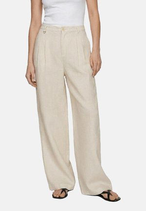 Donna che indossa pantaloni beige a vita alta e gamba larga con pieghe, abbinati a un top bianco senza maniche e sandali neri con cinturini.
