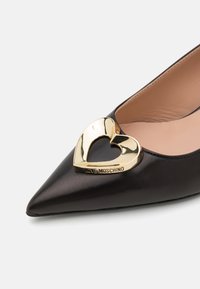 Črne usnjene čevlje s koničastimi prsti, ki imajo velik zlat srčast naglas s predpisom "LOVE MOSCHINO", gladko teksturo in eleganten dizajn.