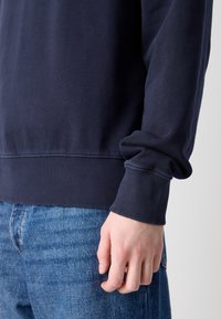 Tmavě modrá mikina s žebrovanými manžetami a lemem, vyrobená z měkké bavlněné směsi; detaily zahrnují hladkou texturu a přiléhavé otvory na zápěstí.