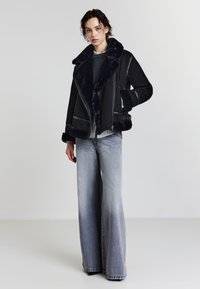 Schwarze Schaffeljacke mit breitem Kragen, Reißverschluss und seitlichem Schnallenverschluss. Kombiniert mit hellgrauen weiten Jeans, die einen kontrastierenden Look erzeugen.