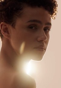 Perfil en primer plano de una persona con cabello corto y rizado. La luz suave crea un resplandor cálido. El tono de piel parece claro, con características faciales sutiles.