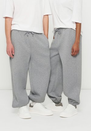 Dve osebi, ki nosita ohlapne sive trenirke z zavezovanjem in bele superge, stojita blizu skupaj na enobarvni beli podlagi.