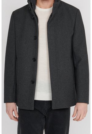 Hombre con abrigo gris oscuro abotonado sobre suéter blanco y pantalones negros de pie contra un fondo liso.