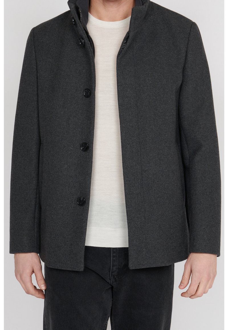 Homme portant un manteau gris foncé boutonné par-dessus un pull blanc et un pantalon noir, debout devant un fond uni.