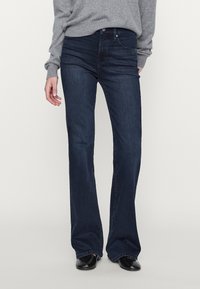 Dark blue, flared jeans with a smooth denim texture, five-pocket design, and subtle stitching details. Worn with a gray sweater and black shoes.