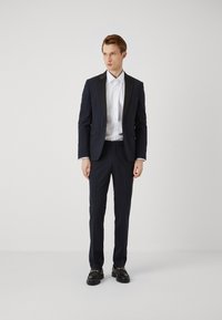 Completo blu con rever nero, pantaloni abbinati e camicia bianca. Le scarpe nere presentano dettagli in oro. Design semplice ed elegante.
