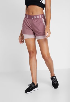 Frau trägt mauvefarbene geschichtete Sportshorts mit "UNDER"-Schriftzug am Bund und schwarze Sportschuhe, posiert vor einfachem Hintergrund.