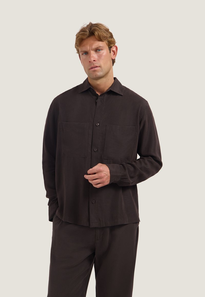 Homme aux cheveux châtain clair portant une chemise marron foncé boutonnée avec deux poches poitrine et un pantalon assorti sur un fond clair uni.