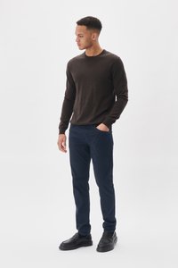 Brązowy sweter z długim rękawem, wykonany z miękkiego materiału, w połączeniu z granatowymi spodniami i czarnymi butami wsuwanymi. Model stoi w neutralnym otoczeniu.