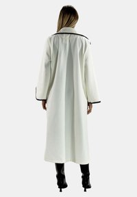 Lang hvid frakke lavet af tekstureret stof med en sort kant langs ærmer og krave. Båret over knæhøje sorte støvler.