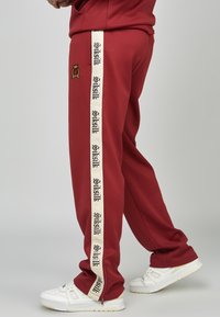 Rote Jogginghose mit geradem Schnitt, elastischem Bund und weißem Seitenstreifen mit wiederholtem schwarzem Text. Ein gesticktes Logo ist sichtbar.