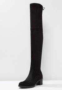 Bottes noires montant jusqu'au genou en faux daim doux, avec un talon bloc épais et une silhouette ajustée. Détails de coutures sur le devant.
