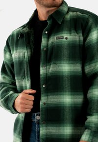 Chemise en flanelle à carreaux verts avec col, boutons-pression et deux poches poitrine, présentant un tissu doux et texturé ainsi qu'une coupe décontractée.