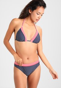 La bikini tummansininen, jossa on valkoisia pilkkuja ja punaisia raidallisia yksityiskohtia. Yläosa on kaula-aukko muotoilulla ja kolmion muotoinen, kun taas alaosa on klassista hipster-tyyliä.