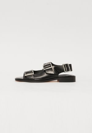 Black leather sandals with rectangular buckles, open toe design, flat sole, and adjustable ankle strap. Smooth texture and minimalistic style.