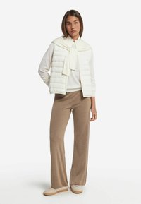 Witte, gewatteerde vest over een crèmekleurige trui, gecombineerd met tan wijdvallende broek en lichte sneakers. Model staat tegen een witte achtergrond.