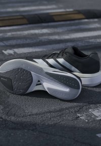 Chaussures de course Adidas noir et blanc sur l'asphalte, une montre la semelle avec les textes visibles "ADIZERO" et "LIGHTSTRIKE".