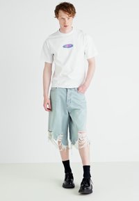 Tricou alb cu un design grafic colorat, asociat cu pantaloni scurți din denim deschis la culoare, lejeri, care au margini destramate și buzunare.