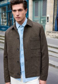 Brun houndstooth jakke med krave-design og forlommer, lagd over en lyseblå skjorte med knapper. Glat tekstur, afslappet pasform.