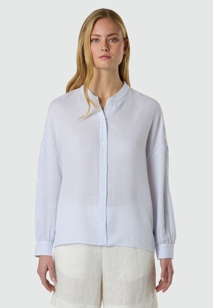 Giovane donna con una camicetta azzurra a righe sottili, con bottoni e maniche lunghe, e pantaloncini bianchi, in piedi davanti a uno sfondo semplice.