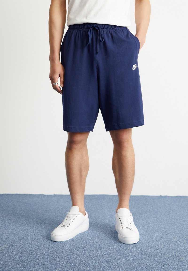 Personne portant un short Nike bleu marine avec taille à cordon et des baskets blanches, debout sur un tapis bleu contre un mur blanc.
