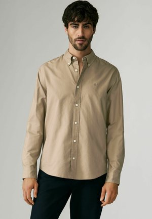 Lys beige skjorte med knapper i bomuld, klassisk krave, lange ærmer og et diskret logo på brystet. Glat tekstur.