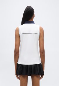 Γυναίκα, εικονιζόμενη από πίσω, φορ ώντας λευκό αμάνικο τοπ με ναυτικό γιακά και μαύρη πλισέ φούστα, πάνω σε απλό λευκό φόντο.