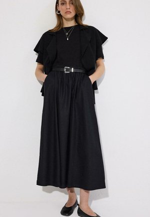 Femme portant un haut noir à manches courtes, une jupe noire à jambes larges avec ceinture, un collier à plusieurs rangs, des chaussures plates noires, debout contre un mur blanc les mains dans les poches.