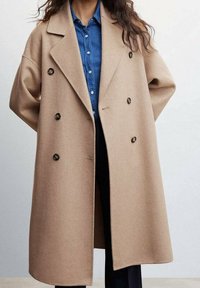 Abrigo cruzado de lana en beige claro. Presenta grandes botones negros, solapas anchas y un corte relajado, superpuesto a una camisa de mezclilla.