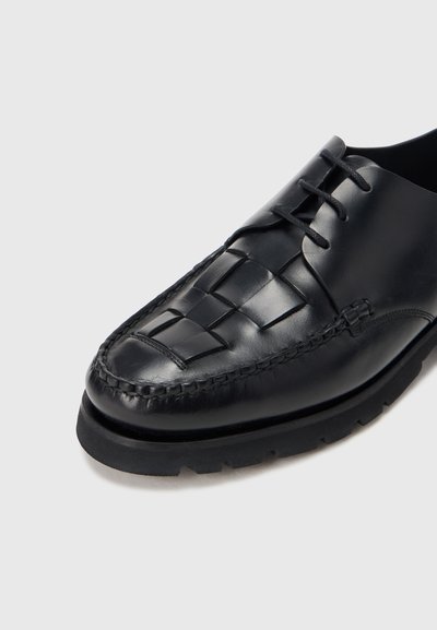 Zwarte leren schoen met geweven patroon op de neus, afgeronde voorkant en contrasterende, gestructureerde zwarte zool. Voorzien van veterontwerp en stikselaccenten.
