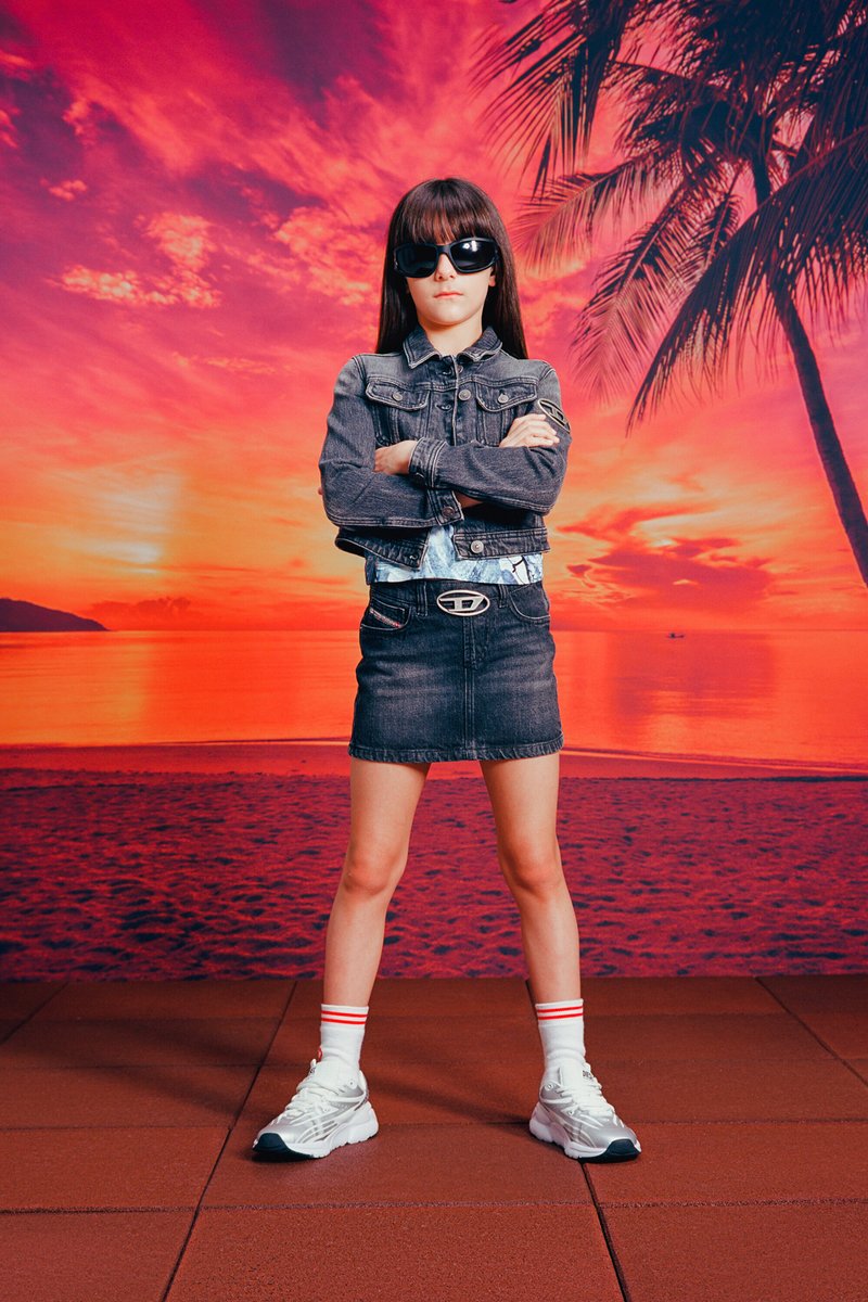 Veste en denim, haut bleu clair, jupe en denim noire, baskets blanches avec des accents gris. Chaussettes rayées blanches et rouges. Lunettes de soleil. Fond de plage.