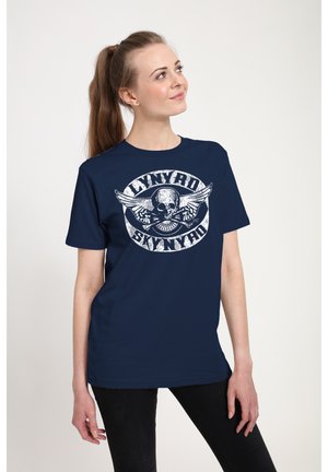 Kvinde med lys hud og brunt hestehale, iført en navyblå Lynyrd Skynyrd T-shirt, kigger let op og mod højre, stående mod en hvid baggrund.