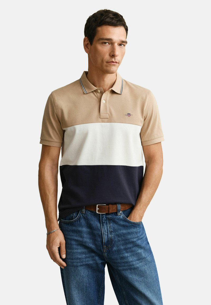 Homme portant un polo à blocs de couleurs beige, blanc et bleu marine avec un jean et une ceinture marron, debout avec une main dans la poche.
