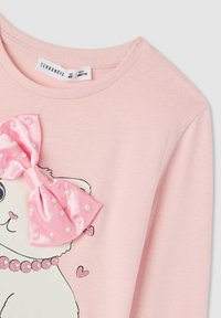 Jasnoróżowa bluzka z długim rękawem z grafiką kota, ozdobiona dużą różową satynową kokardą z białymi perłami oraz błyszczącym kołnierzykiem. Widoczna metka marki.