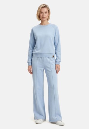 Kobieta stojąca, ubrana w jasnoniebieską bluzę z długim rękawem i pasujące spodnie o szerokich nogawkach z białymi trampkami na jednolitym tle.