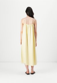 Gul, ärmlös klänning i medellängd med draperad rygg, lätt tyg, avslappnad passform, bärs med svarta sandaler.