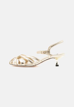 Guld farvet lavhælet sandal med stropper, spænde rundt om anklen og lille kitten heel på en ensartet lys baggrund.