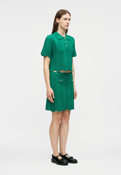 Γυναίκα στέκεται στραμμένη ελαφρώς προς τα δεξιά, φορώντας πράσινο υφασμάτινο κοντομάνικο πόλο μπλουζάκι και ταιριαστή φούστα με κυματιστές άκρες και μαύρα παπούτσια.