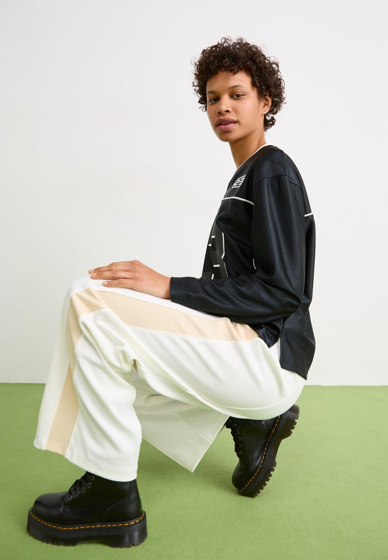 Camisa negra de manga larga con detalles blancos, combinada con pantalones blancos con franjas laterales beige y botas de plataforma negras. Fondo verde.