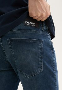 Tummansiniset farkut, joissa on haalistunut tekstuuri, varustettu "DENIM TOM TAILOR" -logotarralla vyötäröllä ja tavallisilla takataskuilla.