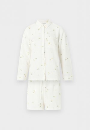 Fehér hosszú ujjú ing kis sárga virágmintával és hozzáillő fehér rövidnadrággal egyszínű háttéren.