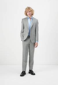 Gray tailored suit with a notched lapel, paired with a light blue shirt. Model stands confidently, wearing black shoes. Smooth texture throughout.