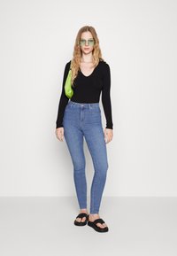 Fekete, bordázott, hosszú ujjú felső, magas derekú kék farmer, fekete papucs és zöld táska. A modellen zöld árnyalatú napszemüveg van.