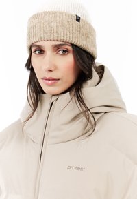 Beige geïsoleerde jas met een hoge kraag en ritsen, gecombineerd met een tan en crèmekleurige gebreide beanie met een label.