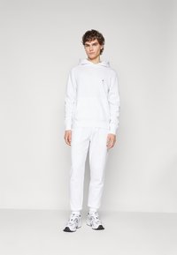 Sweatshirt blanc en coton avec poche avant et poignets côtelés, associé à un pantalon de jogging blanc. Le mannequin porte des baskets blanches et noires. Design simple et minimal.