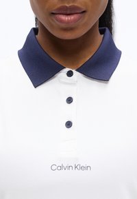 Εγγύς πλάνο ενός ατόμου που φοράει λευκό πόλο μπλουζάκι Calvin Klein με γιακά σε ναυτικό μπλε χρώμα και τρία κουμπιά στο ίδιο χρώμα.