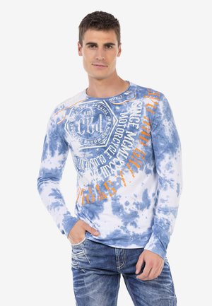 Long-sleeve shirt with blue and white tie-dye pattern, featuring orange graphic text and a round neckline. Worn with blue jeans.