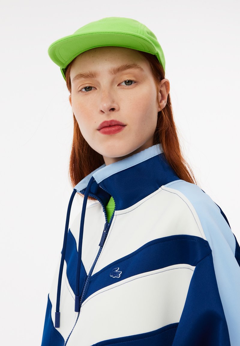 Chaqueta azul y blanca con cremallera y diseño en chevron, que presenta un cuello alto y cordones. Combinada con una gorra verde. Textura suave.